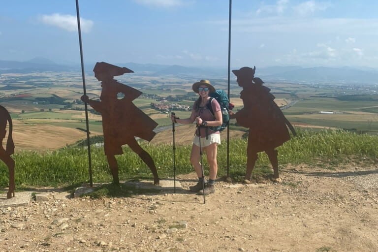 Pilgrim on the Camino de Santiago smiling at the top of a mountain with iron statues of pilgrims behind her.