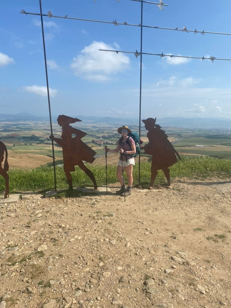Mikala on the Camino de Santiago smiling at the top of a mountain with iron statues of pilgrims behind her.