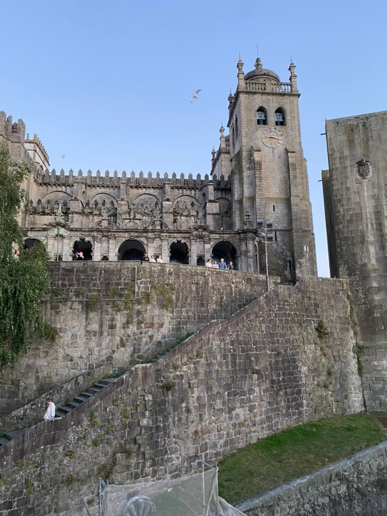 Outside of the Porto Cathedral in Porto, Portugal. The cathedral sits high on top of a hill with people looking over a stone wall and a staircase leading down to the street below.