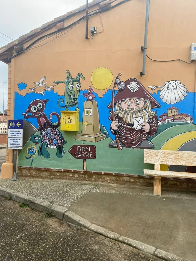 Wall mural depicting an ancient pilgrim holding an envelope to put into the real mailbox and surrounded by a Camino trail marker and animals in the mural