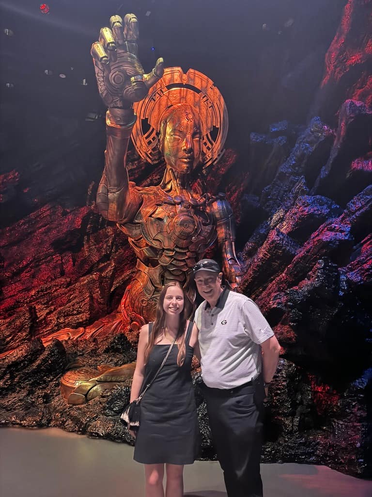 Mikala and her dad standing smiling in front of a massive robot animatronic behind them at Wētā Workshop Unleashed in Auckland.