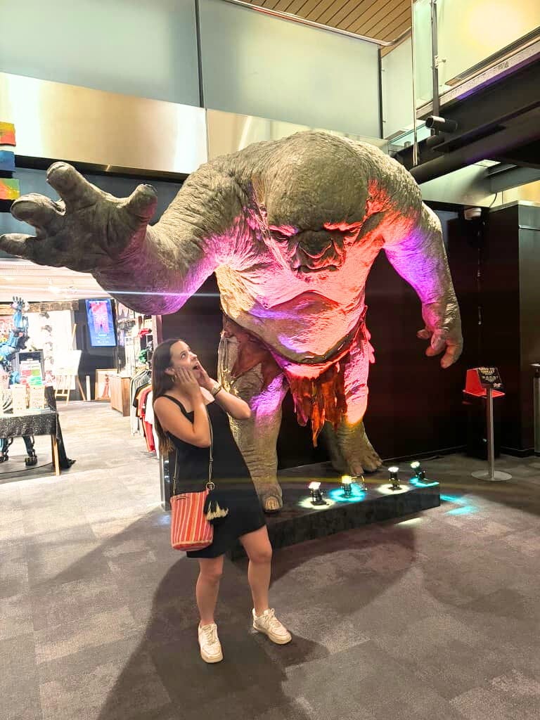Mikala standing pretending to be scared in front of a massive The Lord of the Rings troll statue at Wētā Cave at Wētā Workshop Unleashed in Auckland.