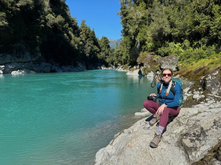 Mikala sitting smiling on a rock in front of Hokitika Gorge in New Zealand