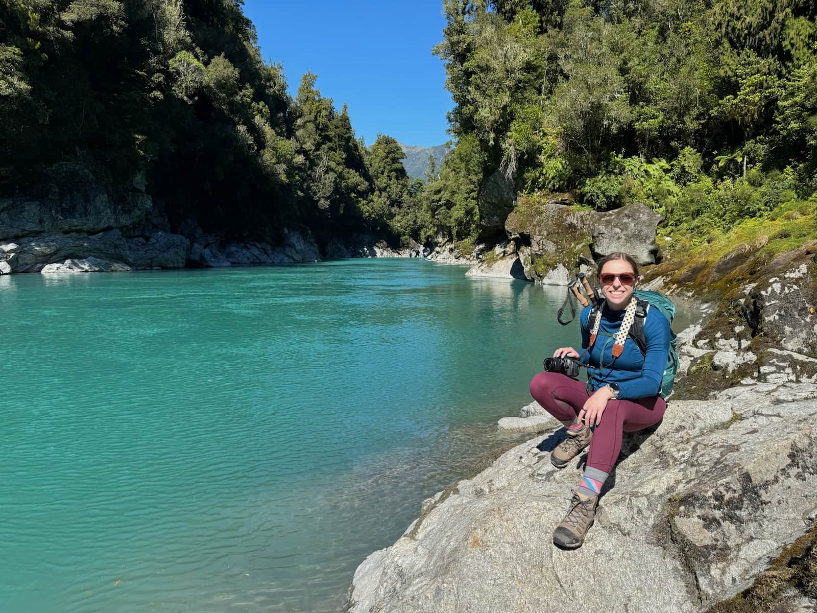 Mikala sitting smiling on a rock in front of Hokitika Gorge in New Zealand
