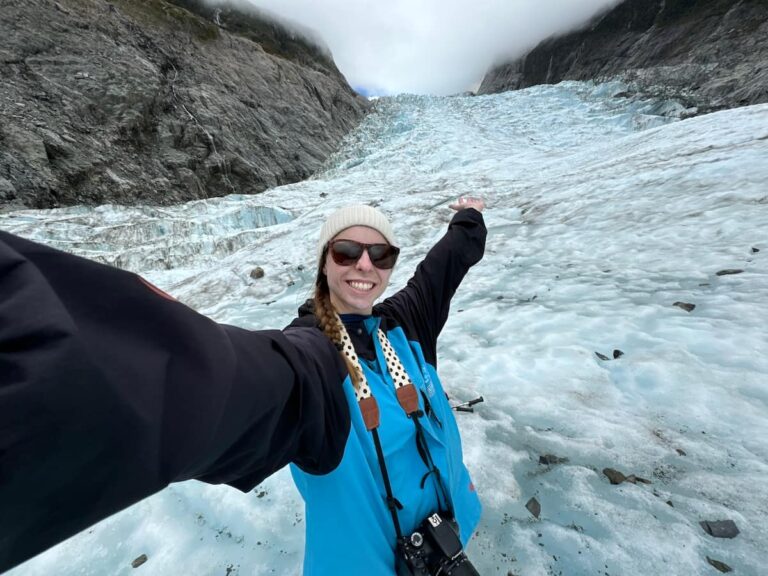 Mikala taking a selfie with her hand up in the air in front of Fox Glacier while on a heli hiking tour in New Zealand.