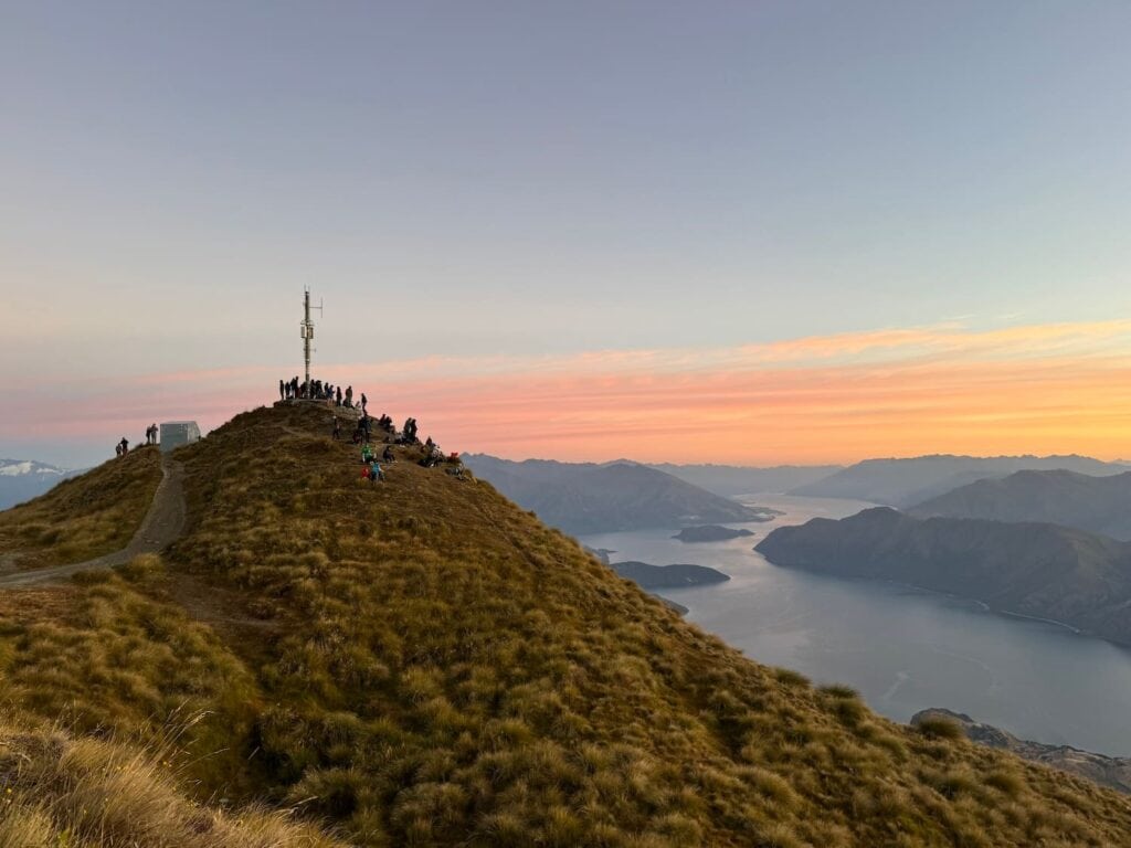 Group of people crowded on a mountain summit with a cellular antenna on Roy's Peak at sunrise.