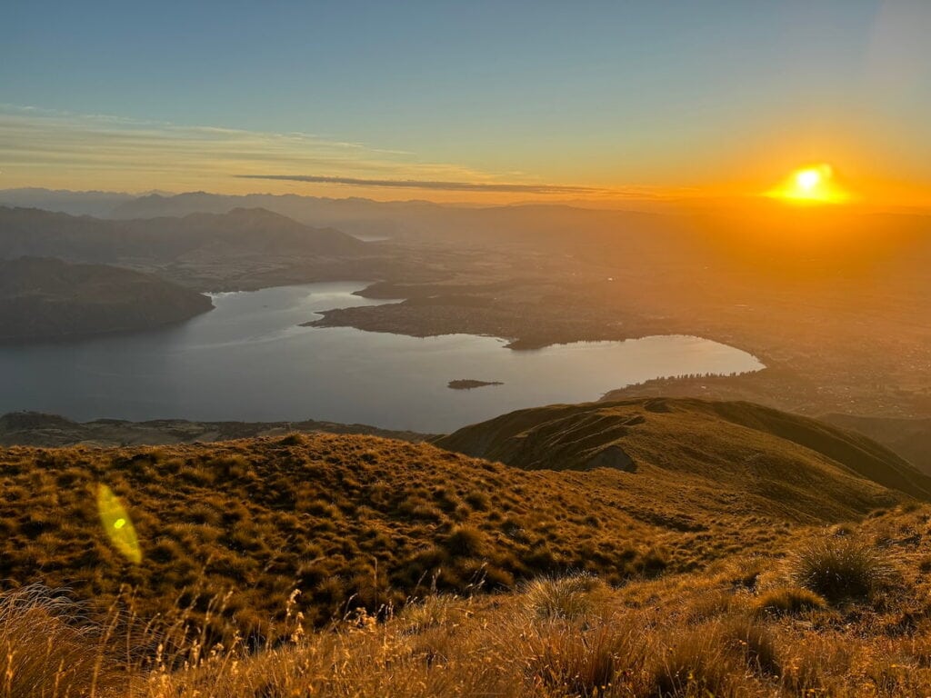 Bright orange sun appearing over the horizon over Wānaka and Lake Wānaka, taken from the Roy's Peak summit.