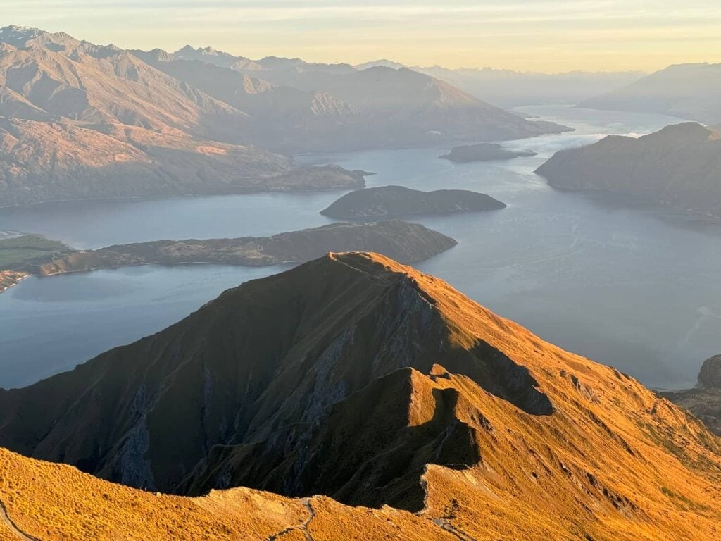 Mountain with trails to Roy's Peak bathed in golden light from sunrise with Lake Wānaka and other mountains in the background.