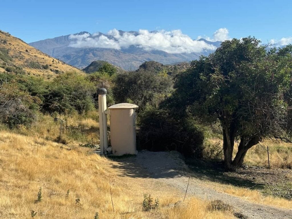 Drop toilet surrounded by grass and trees with a mountain in the background. This is the toilet in the parking lot at the Roy's Peak Track trailhead.