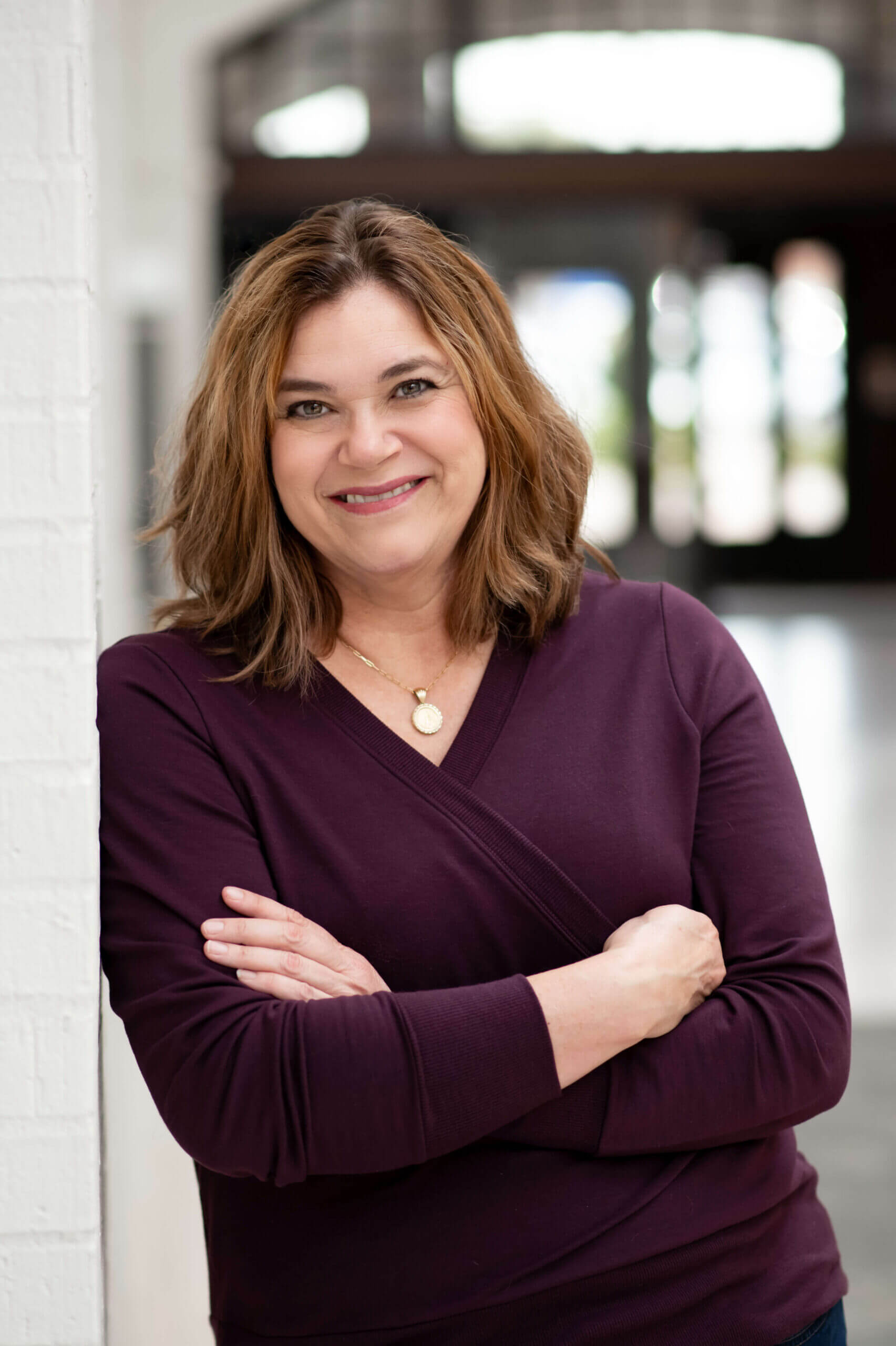 A person with shoulder-length brown hair, wearing a purple top and a necklace, stands smiling with arms crossed in a bright indoor space. Solve your lice problems with Lice Nannies USA.