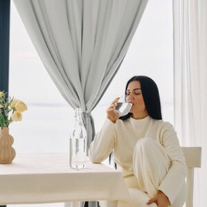 A woman with long brown hair, dressed in a cream tracksuit, sat at a kitchen table drinking a glass of water before trying out one of our 5 minute morning journaling routines