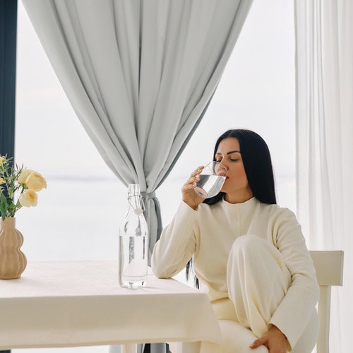 A woman with long brown hair, dressed in a cream tracksuit, sat at a kitchen table drinking a glass of water before trying out one of our 5 minute morning journaling routines
