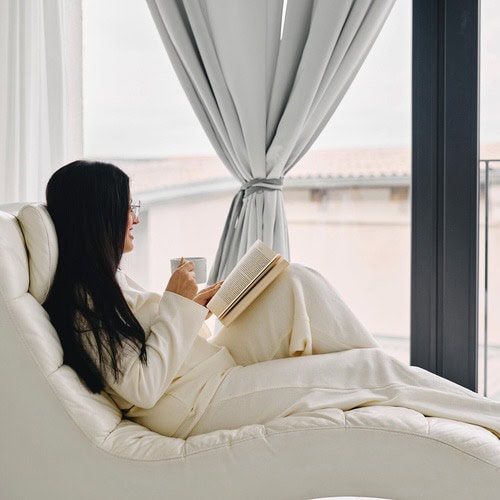 A woman with long brown hair, dressed in a cream tracksuit, sat in a cream arm chair writing her morning journal pages