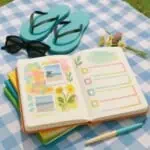 A photo of an open bullet journal set up to use our summer journal ideas. On the page are some doodles and boxes to write in in blue, yellow and orange, a photo of a plant and some yellow washi tape. The journal is on a picnic blanket next to some flip flops