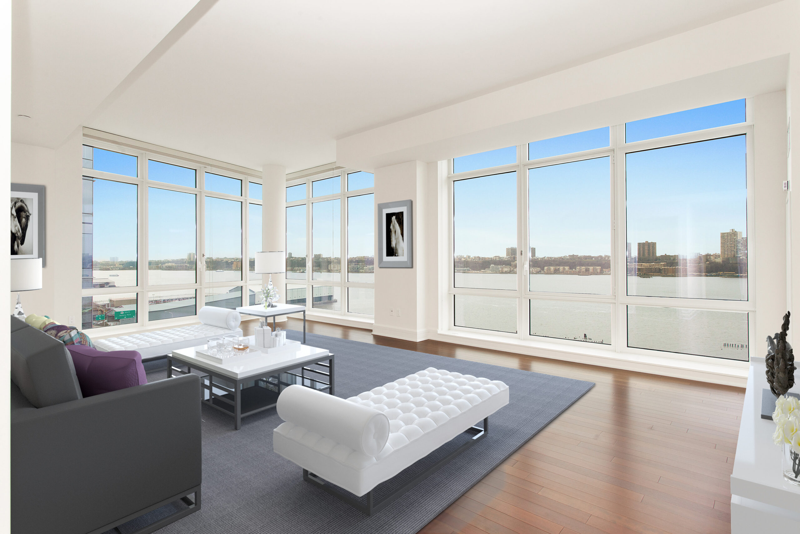 Bright modern living room at The Aldyn with floor-to-ceiling windows overlooking a river, featuring minimalist white furniture, a gray rug, and hardwood floors. Artwork and decor add subtle accents to the spacious, open layout.