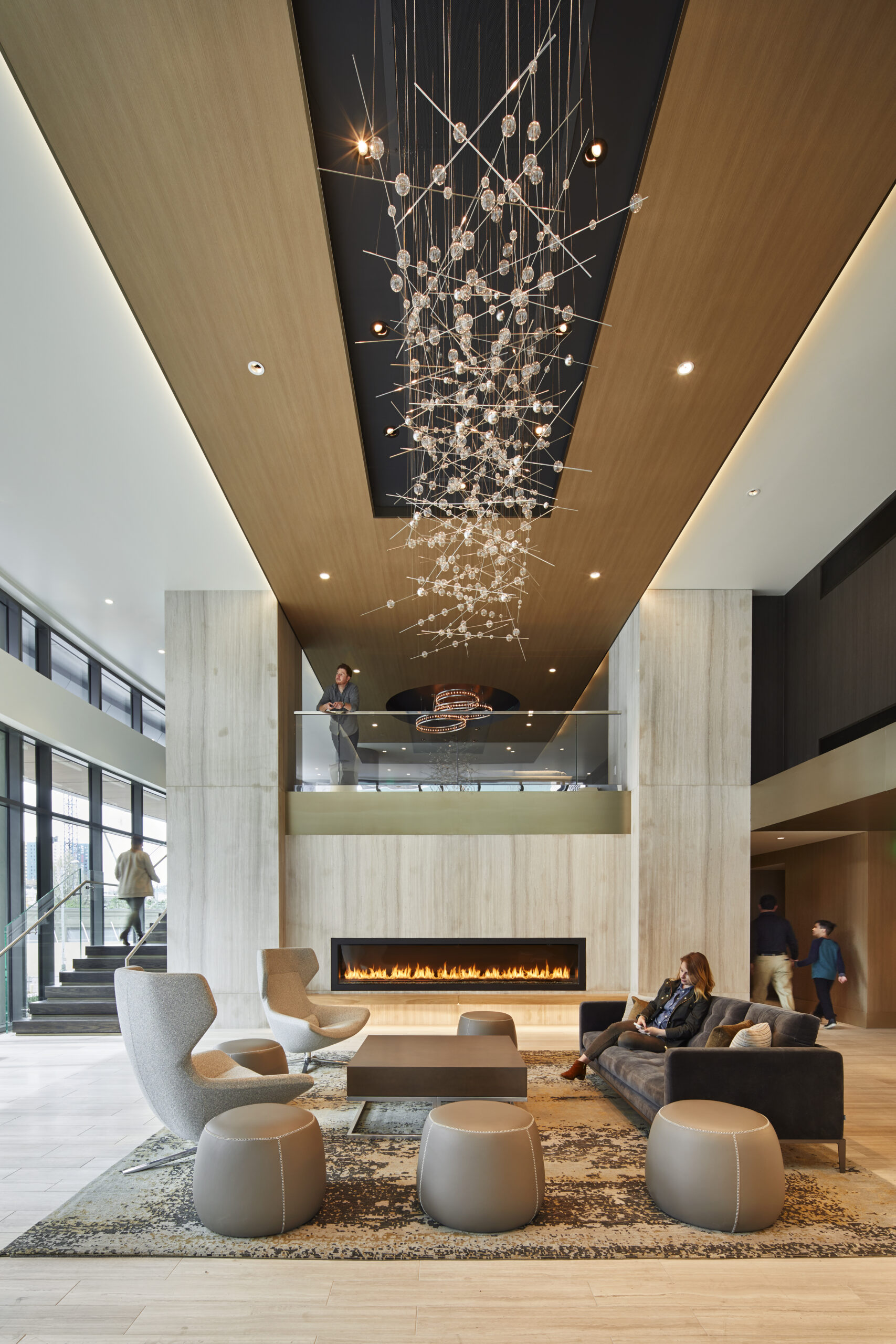 Modern hotel lobby with tall ceilings, a large Stratus chandelier, contemporary furniture, a long fireplace, and people walking or sitting in a relaxed atmosphere.