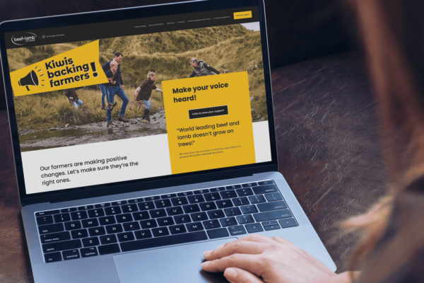 A person views a Beef & Lamb website on a laptop, displaying a banner with "Kiwis backing farmers!" and an image of people working in a field. The site encourages support for farmers, with text urging readers to "Make your voice heard!" and mentions "world leading beef and lamb.