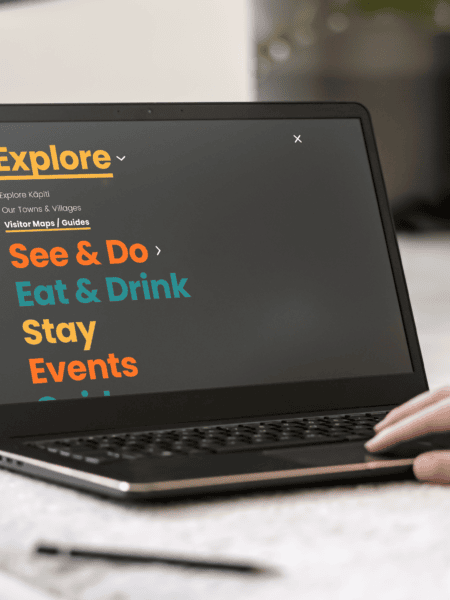 A person using a laptop displaying a Kāpiti regional tourism website section titled "Explore" with categories: See & Do, Eat & Drink, Stay, Events. The text is brightly colored on a dark screen. The scene includes a carpet and a plant in the background.