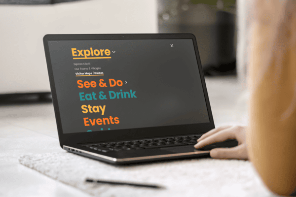 A person using a laptop displaying a Kāpiti regional tourism website section titled "Explore" with categories: See & Do, Eat & Drink, Stay, Events. The text is brightly colored on a dark screen. The scene includes a carpet and a plant in the background.