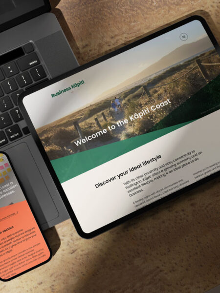 A tablet displaying a welcome screen for the Kapiti Coast Business website with a scenic image and text, placed on a wooden table next to a smartphone showing an orange and green app screen, and a partially visible laptop keyboard.