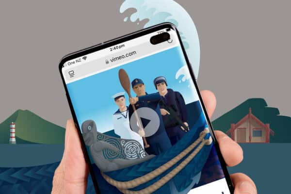 A hand holding a smartphone displaying an illustration of three sailors in an animated video on vimeo.com. The background shows a large wave, a lighthouse, and green hills. A carved grey figurehead of a waka and rope are visible at the bottom of the phone screen.