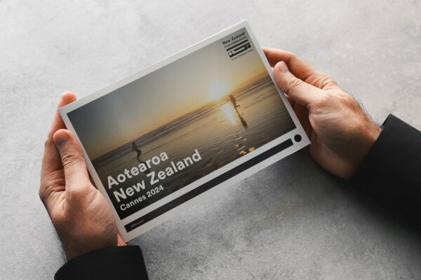 A person holds a brochure featuring a beach scene with two people at sunset. The text reads "Aotearoa New Zealand Cannes 2024" with a New Zealand logo in the corner.