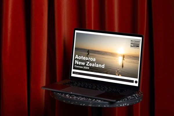 A laptop sits on a round table in front of red curtains. The screen displays an image of people walking along a beach at sunset with text reading "Aotearoa New Zealand Cannes 2024" and a logo for New Zealand Film Commission.