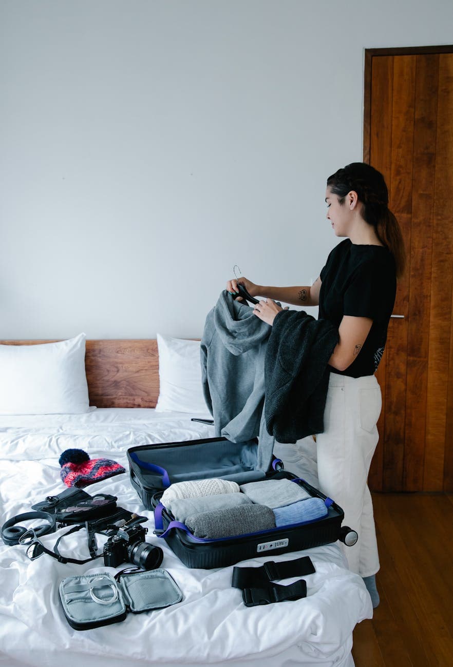 a woman in black t shirt packing clothes beside a bed with suitcase