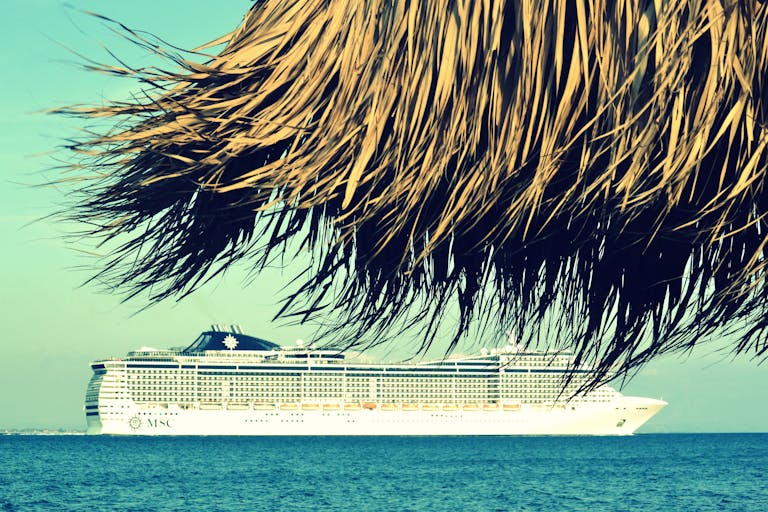 A cruise ship sails past a tropical beach in Katakolo, Greece, under clear skies.