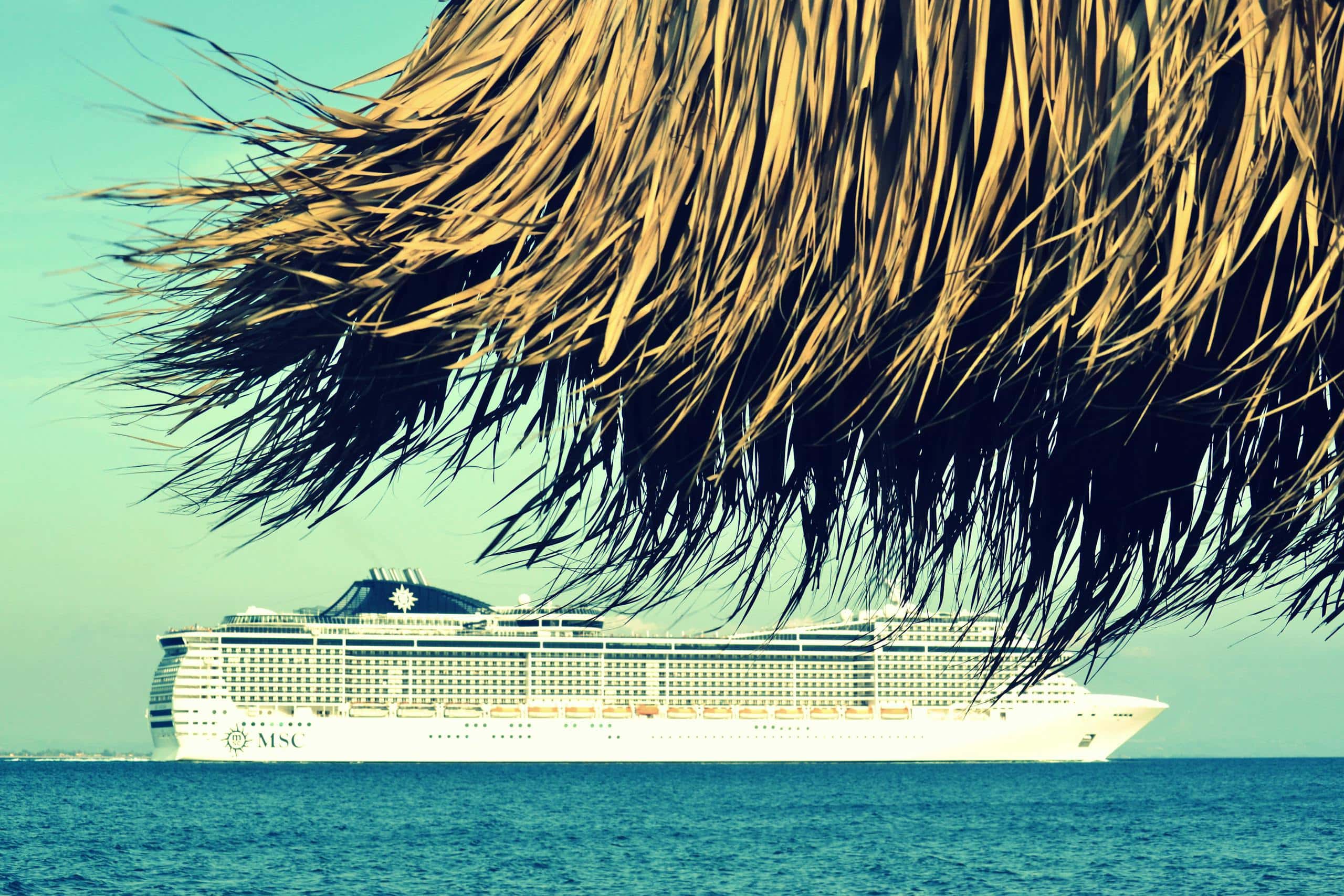 A cruise ship sails past a tropical beach in Katakolo, Greece, under clear skies.