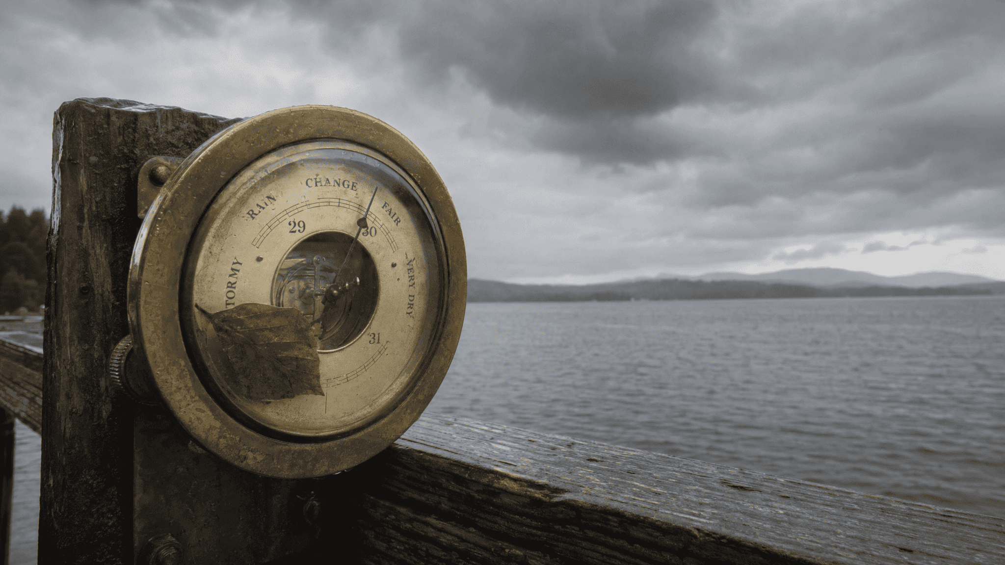 Brass analog barometer on a weathered dock rail under an overcast sky over a wide grey lake