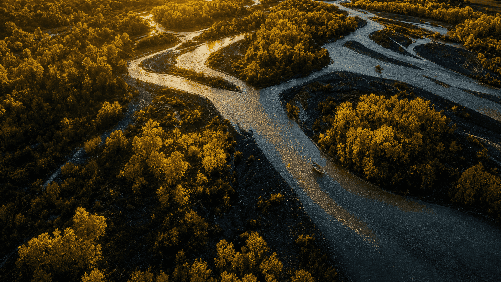 Aerial view at golden hour of a braided mountain river through cottonwood bottomland, a single wooden drift boat anchored mid-channel, no landmarks visible