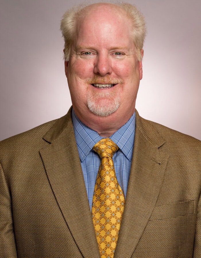 Smiling male business professional wearing a brown jacket and gold patterned tie.