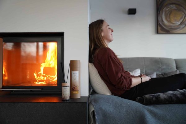 Frau entspannt sich auf dem Sofa neben einem brennenden Kamin und AlpenXund Raumduft – Wohlfühlatmosphäre zu Hause.