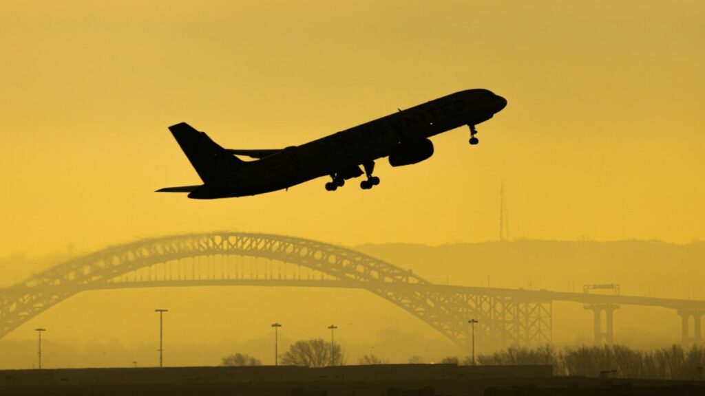 Aereo in decollo al tramonto con ponte e paesaggio urbano sullo sfondo.