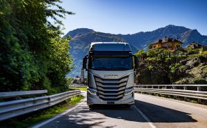 Camion Iveco S-Way grigio in marcia su una strada panoramica tra le montagne, con una casa isolata su una collina e una chiesa sullo sfondo.