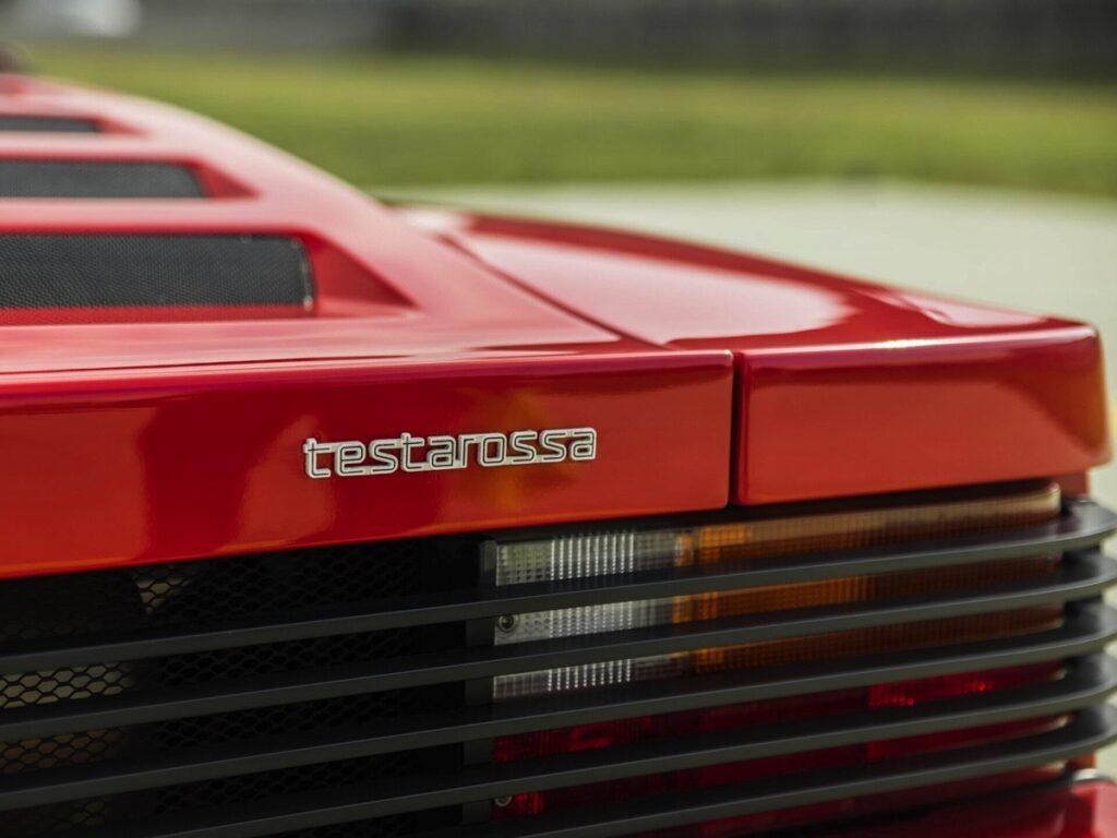Dettaglio posteriore Ferrari Testarossa rossa con logo originale e gruppo ottico, ripresa ravvicinata su sfondo sfocato