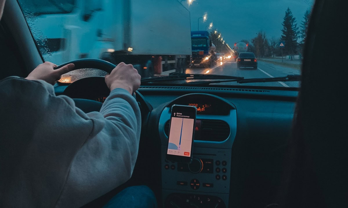 Vista interna dell’auto con conducente e navigatore acceso durante la guida serale sotto la pioggia.