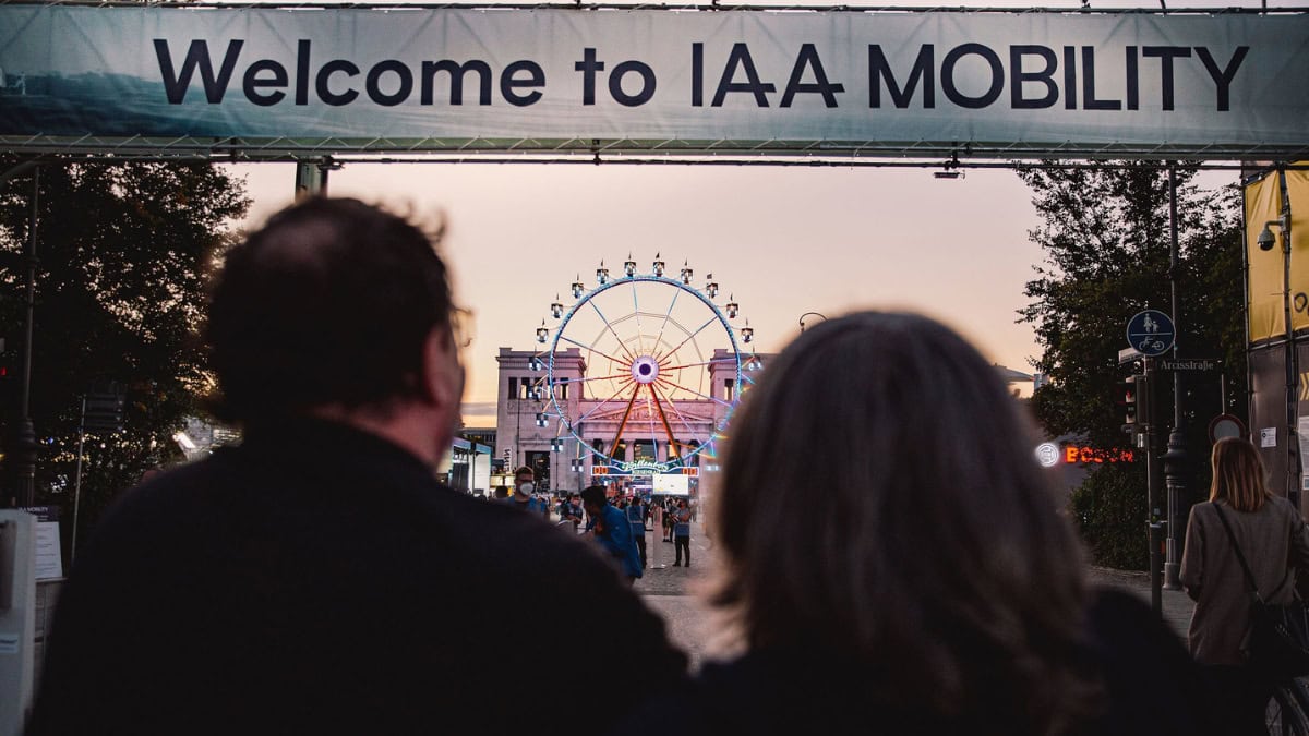 Ingresso alla fiera IAA MOBILITY con ruota panoramica in background e visitatori in prima piano, in un'atmosfera serale di evento automotive e mobilità sostenibile