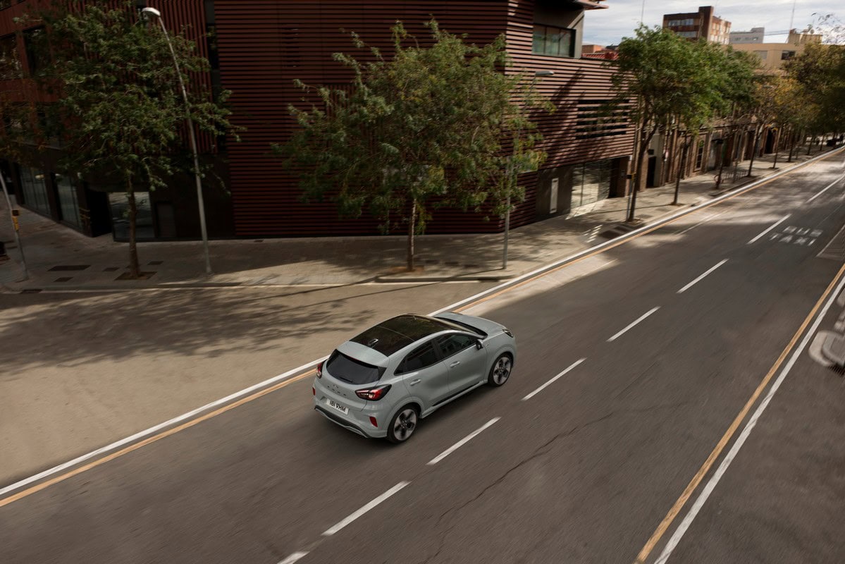 Ford Puma grigia vista posteriore a 3/4 dall’alto, in marcia su strada urbana alberata con edifici moderni