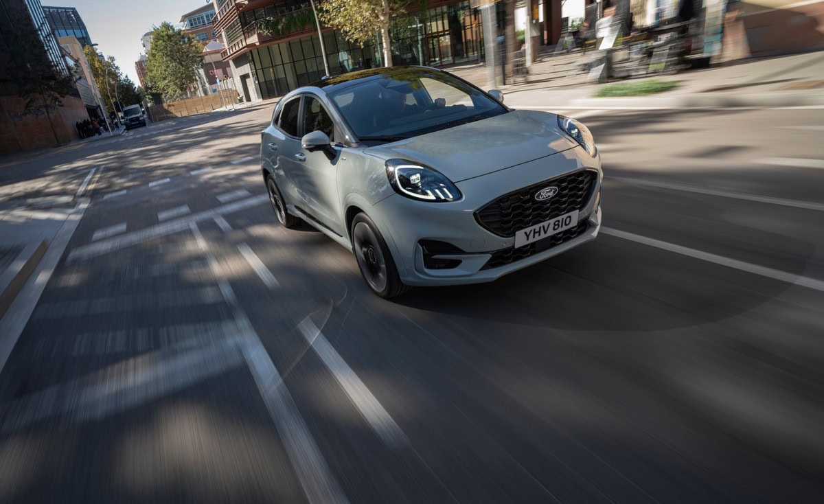 Ford Puma grigia vista frontale in movimento su strada urbana, ripresa a 3/4 con sfondo cittadino sfocato