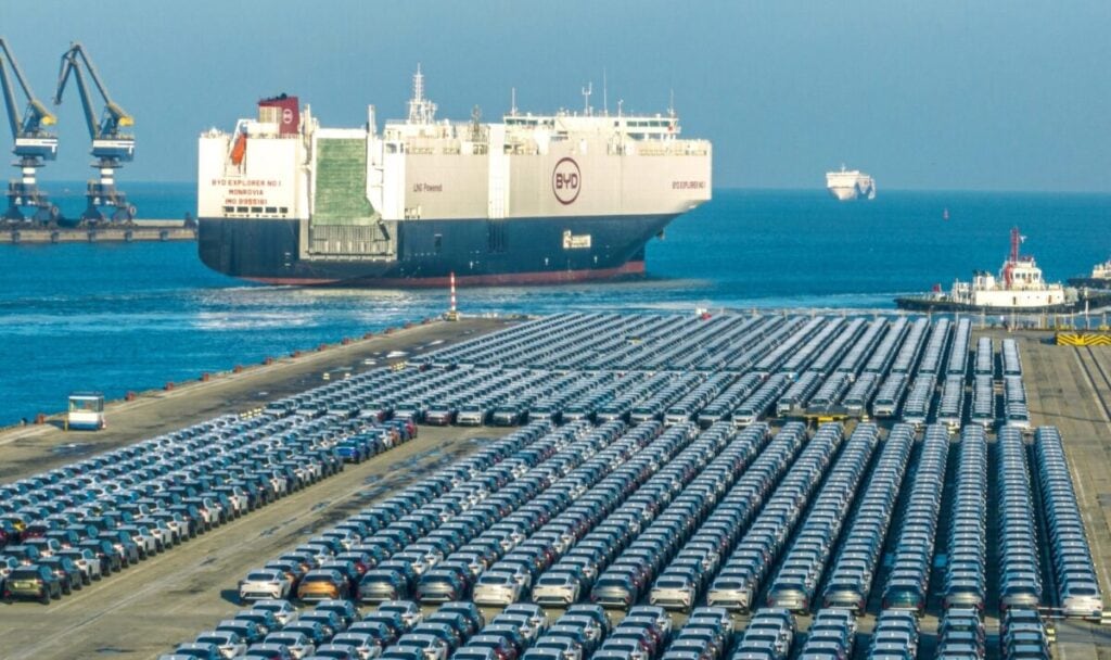 Parco auto in porto con grande nave cargo BYD e container, trasporto merci marittimo, terminal portuale, logistica e spedizioni internazionali