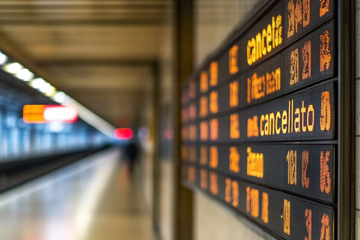 Stazione ferroviaria con tabellone elettronico orari treni in partenza e cancellati, ambiente urbano, trasporto pubblico.