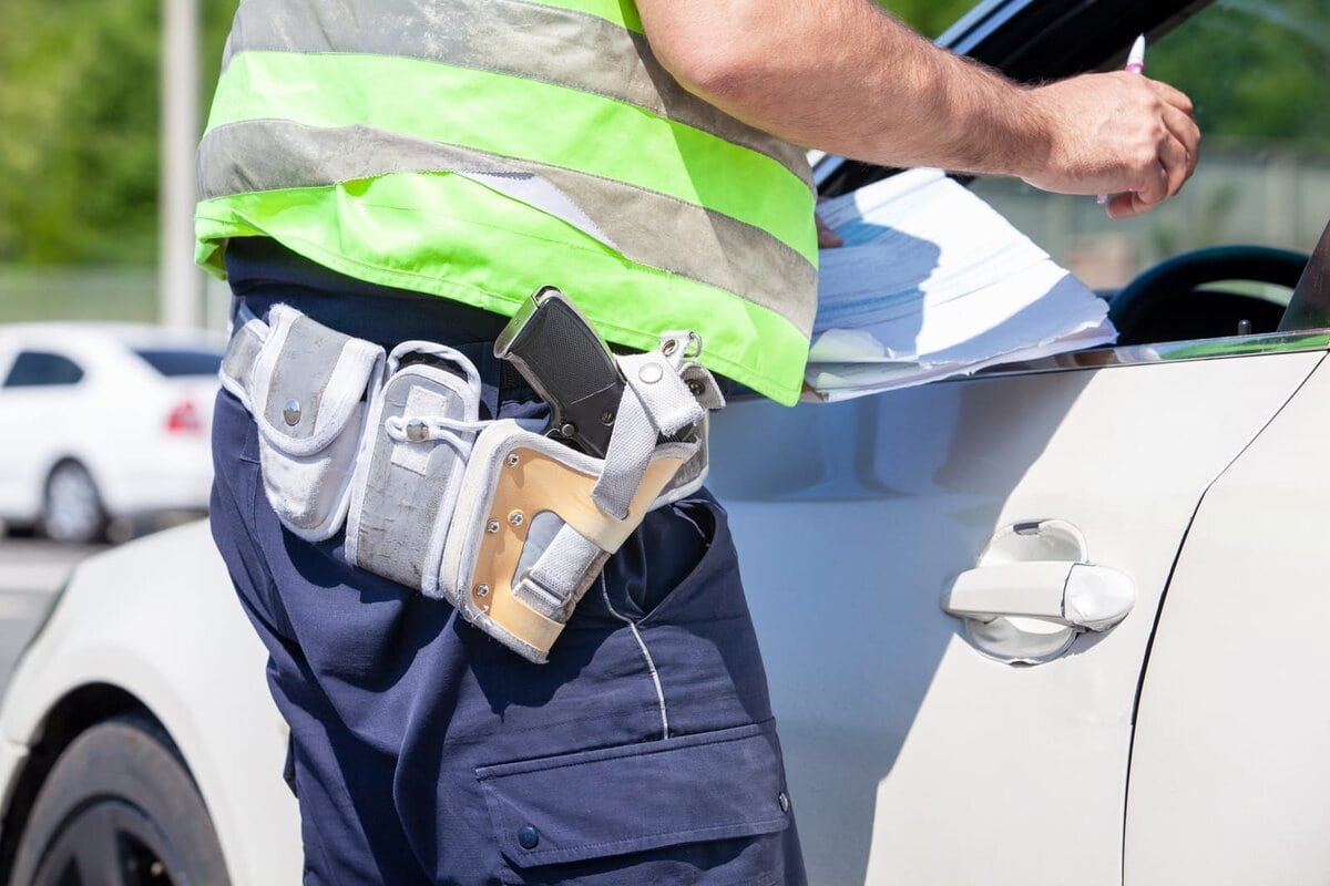 Agente di polizia con fondina e gilet riflettente mentre compila una multa appoggiato a un'auto bianca durante un controllo stradale autovelox.