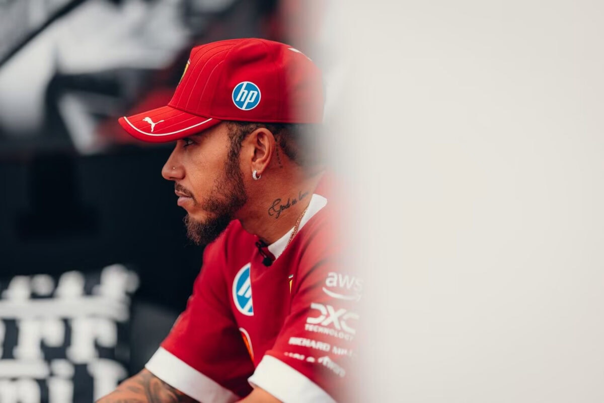 Lewis Hamilton con cappellino e maglia Ferrari F1 guarda concentrato verso sinistra, in un'inquadratura laterale con sfondo sfocato.
