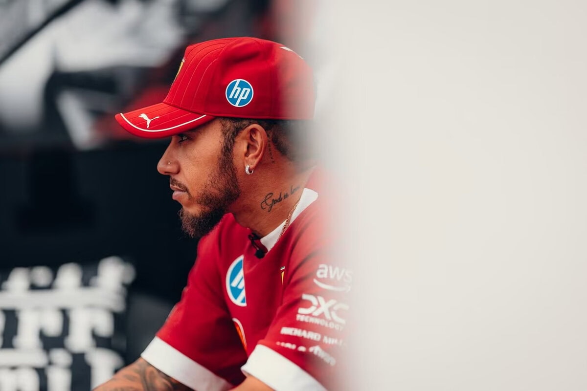 Lewis Hamilton con cappellino e maglia Ferrari F1 guarda concentrato verso sinistra, in un'inquadratura laterale con sfondo sfocato.