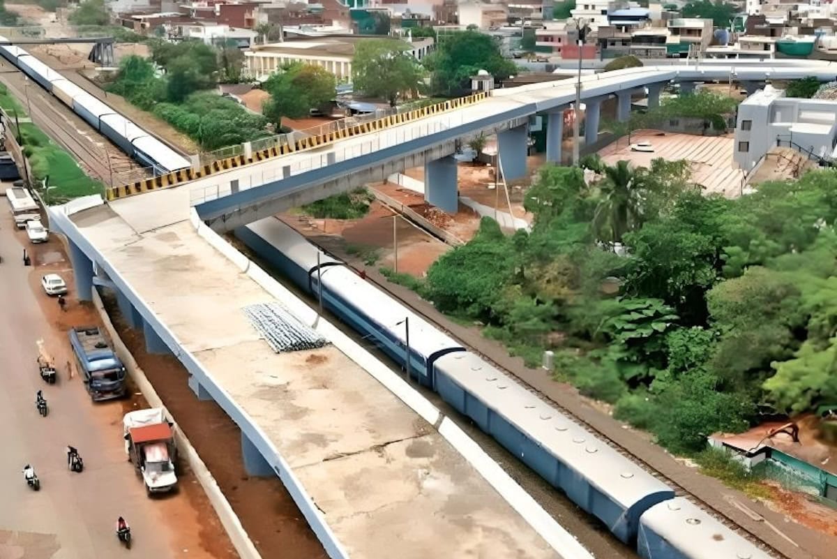 Strada sopraelevata in costruzione a Bhopal sopra la linea ferroviaria in area urbana, con traffico leggero e vegetazione ai margini.