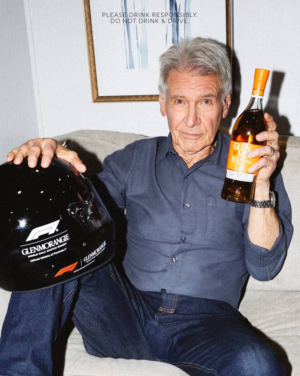 Harrison Ford con casco F1 Glenmorangie e bottiglia di whisky in mano, seduto su divano con espressione rilassata