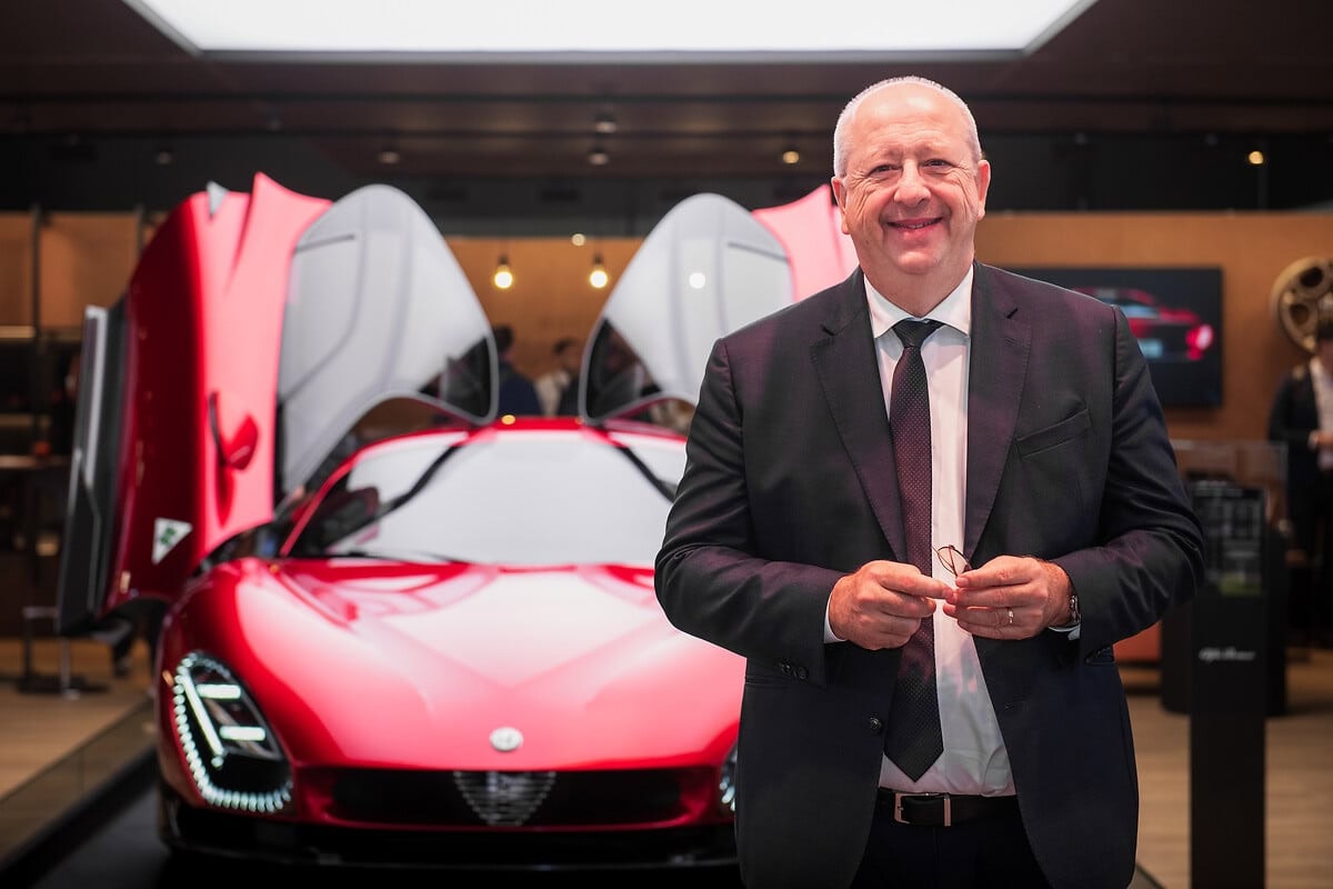 Jean-Philippe Imparato in abito scuro davanti a supercar Alfa Romeo rossa con portiere ad ali di gabbiano aperte.