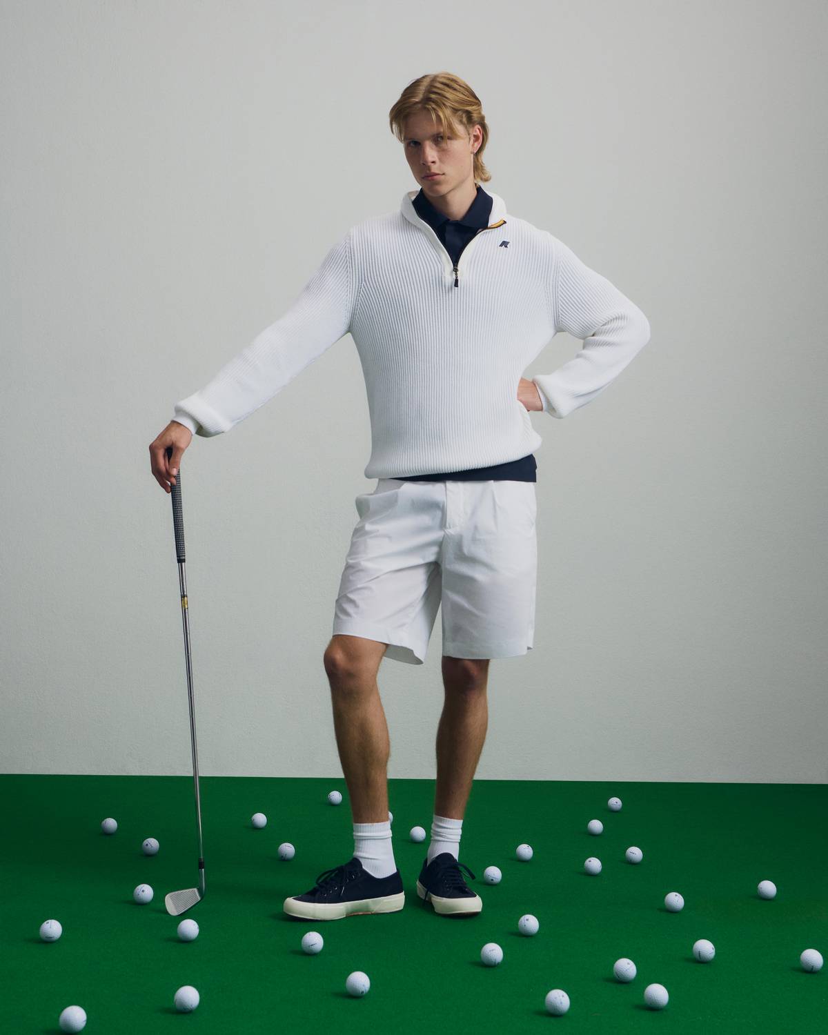 Giovane golfista in maglione bianco a mezza zip, polo blu e pantaloncini, su prato verde